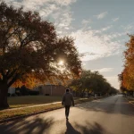 Person strolling down a tree-lined street with oak trees in vibrant fall foliage and dappled sunlight on the pavement