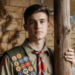 Brandon Barrett Eagle Scout standing proudly before wooden scouting post with backpack full of merit badges map background