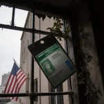 Green card visa holder hangs from gate with dust and vines covering the metal and a faded American flag behind