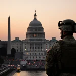 National Guard soldier standing watch on a raised platform with the U.S. Capitol at sunset and the Washington Monument in bac