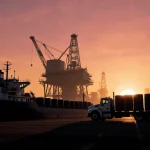 Tanker ship unloading oil barrels onto truck with sunset sky and looming oil rig