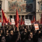 Protesters chanting with signs and banners beside a grand building with a faded communist mural surrounded by red flags
