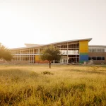 Field expands with mature trees and a modern school under construction bathed in warm sunlight.