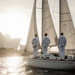 Two French sailors standing back-to-back at helm of 35‑ft yacht with Opera House in foggy dawn background
