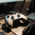 Sleek futuristic gaming controller glimmers on cluttered desk with tangled wires and gaming gloves nearby.