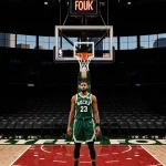 Giannis Antetokounmpo staring at rim with limp net and empty seats in dimly lit Milwaukee Bucks court