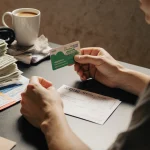 Person sits holding an unopened gift card on a cluttered desk with a half‑full coffee mug and stacks of cash envelopes