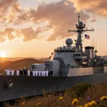 Golden Fleet flagship battleship glimmers against a sunset-lit Texas Hill Country backdrop with a crowd of Texans on deck.