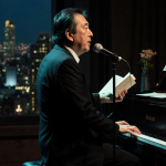 Haruki Murakami stands at microphone with glowing piano keys and cityscape window behind