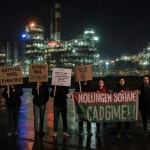 Hungarian residents protesting with banners near a battery factory and neon lights reflecting on wet pavement