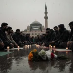 Mourners seated on ground with candles and prayer mats in rain near mosque minaret