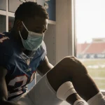 Quinshon Judkins lies on stretcher with leg bandaged and mask over face and light from window and locker room medical team.