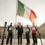 Four Italian athletes stand proudly with the Italian flag held high and the presidential palace backdrop with skis and snowbo