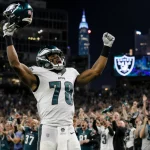 Jalen Hurts stands triumphantly holding his helmet high with cheering Eagles fans and skyline reflected in stadium lights.