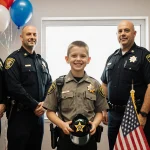Young Jaxson proudly holding a toy sheriff hat with a deputy badge while standing before a Texas flag and balloons