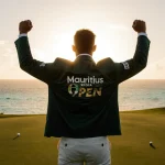 Jayden Schaper triumphantly raising his arms with sunset gold light on a lush golf course and turquoise waters nearby