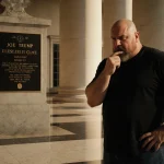 Joe Rogan looks sternly at a Trump plaque on the White House Colonnade with polished marble floor reflecting grandeur.