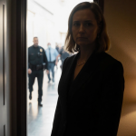 Judge Hannah Dugan standing alone in a dim hallway with a slightly ajar back door and faint agent shadows.