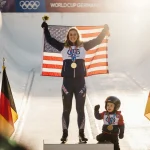Kaillie Humphries Armbruster holds her gold medal aloft with her child beside her and the U.S. and German flags behind her