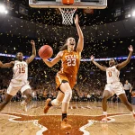 Kyla Oldacre dribbling toward the hoop with confetti swirling and teammates cheering in a packed Texas stadium.