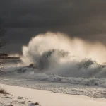Massive wave crashing onto Lake Superior shore with white foam and dark gray sky and bare trees swaying