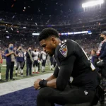 Lamar Jackson sits on bench clutching his back with frustration while Patriots celebrate behind him under stadium lights