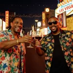 UB40 members toast glasses of Red Red Wine with neon lights and countdown clocks on the Las Vegas Strip at dusk.