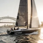 LawConnect yacht cutting through waters with billowing sails and Sydney Harbour Bridge at sunrise.