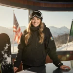 Lindsey Jacobellis addressing camera NBC booth and snowboarding gear beside her Italian winter backdrop