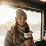 Lindsey Jacobellis stands with microphone under Italian sunlight beside Olympic medals on shelf