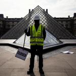 Worker holding picket sign with Louvre pyramid and Closed sign in background and protest banners fluttering