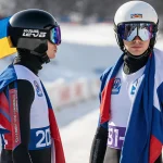 Ukrainian and Russian lugers stand side by side at the World Cup starting line with flags on helmets in snowy background