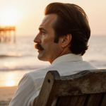 Anthony Geary as Luke Spencer sits on a worn wooden beach chair with a golden sunset and a blurred hospital set behind him