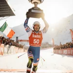 Marco Odermatt crosses the finish line with his 50th World Cup trophy and fans cheering under golden hour light