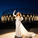 Mariah Carey raising an arm with a sparkling white gown and Italian flag and Olympic lights behind the San Siro stadium.