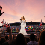 Mariah Carey sings atop San Siro steps with microphone and sparkling gown against sunset sky and twinkling lights