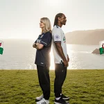 Martha Stewart and Snoop Dogg standing back‑to‑back looking at Swansea stadium with green grass and Welsh coast.