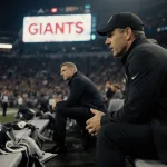 Maxx Crosby sits on Raiders bench with Coach Pete Carroll explaining bench decision and a blurred Giants logo looming behind.