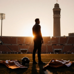 Michael Taaffe standing alone on the football field with gear and Longhorn scarves as the Tower of Texas looms in sunset.