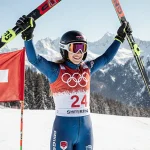 Mikaela Shiffrin stands triumphantly at finish line with skis pointing skyward and Swiss flag waving behind snow-capped mount