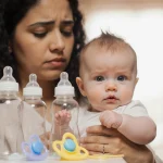 Mother holding infant with concerned expression near shelf of ByHeart formula and empty baby bottles.