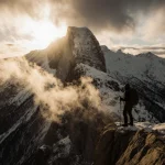 Hiker silhouette standing on Mt. Baldy snow‑capped cliff with rope and sunrise light