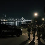 National Guard troops standing at attention with black SUVs and silver Mississippi River in a nighttime New Orleans street.
