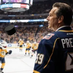 Nick Saban beside hockey puck with Nashville Predators jersey draped over shoulder as Gnash mascot looks up glass in stadium.