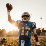 Nico Iamaleava stands proudly on UCLA field at sunset holding football aloft with fallen leaves and warm light.