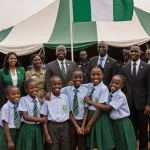 Smiling children in school uniforms holding hands with Nigerian flag behind tent and security guards watching
