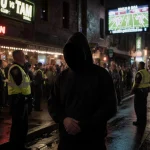 Lone figure in black hoodie raises hands in front of a chaotic bar with police flashing lights and neon glow on wet pavement