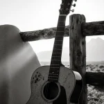 Old worn guitar resting against a weathered wooden fence with long shadows and a hint of distant mountains.
