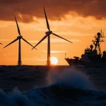 Wind turbines standing still with navy ship hull waves below and radar antenna rising beyond sunset sky
