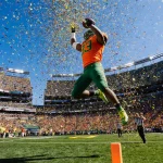 Oregon Ducks player leaps with football while confetti explodes and orange fans cheer